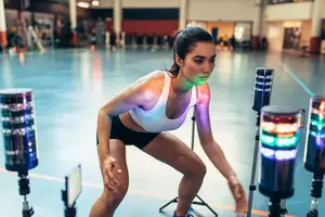 A woman in a gym, preparing for an exercise, with a focus on her stance and the colorful lights in the background.