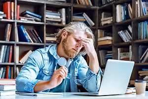 A man sitting in front of a laptop with his eyes closed and hand on his forehead