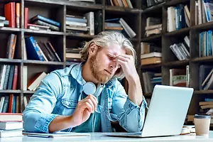 A man sitting in front of a laptop with his eyes closed and hand on his forehead