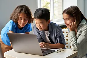 Three children are gathered around a laptop on a table, with one child looking at the screen, another child is holding a pencil, and the third child is listening attentively.