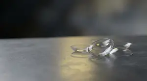 Three clear glass contact lenses sit on a metallic surface in the foreground.