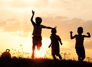 Three children jump together on a grassy field as the sun sets in the background