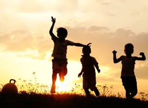 Three children jump together on a grassy field as the sun sets in the background