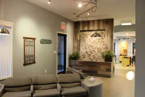 An interior view of a modern office or waiting area with gray walls, wooden elements, and a cozy seating arrangement, featuring a brick wall, a wooden reception desk, and a ceiling with track lighting and lamps.