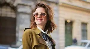 A woman is standing on a street corner wearing sunglasses, a green coat, and a white collared shirt with a blue tie. Behind her is a building with a green door and window, and a person walking on the sidewalk. A car is parked on the side of the road. The background is blurry.