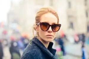 A woman wearing sunglasses and a blue coat is standing in a busy street with buildings and people in the background.