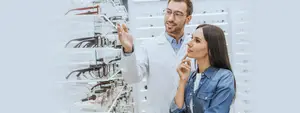A man in a lab coat is pointing at glasses on a shelf while a woman looks on.