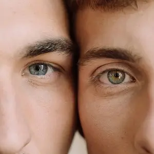 Close-up view of two men's faces, one with brown eyes and the other with blue eyes, they are standing close to each other, probably in a studio