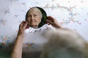 An elderly woman is looking at her reflection in a mirror with a floral pattern on the wall behind her.