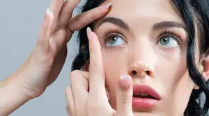 A close-up of a woman inserting contact lenses into her eye while another person helps her.