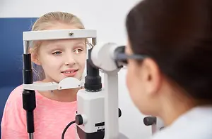A girl with blonde hair and blue eyes is looking at a doctor while sitting on a chair and wearing a pink shirt.