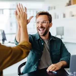 A man in a green jacket is smiling and giving a high-five to another person in an office setting.