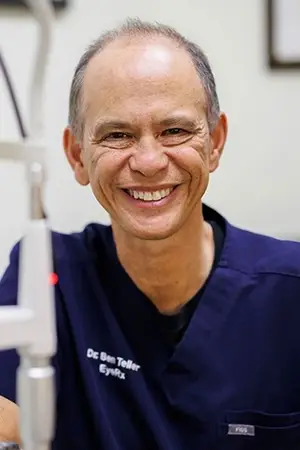 A smiling man wearing a blue shirt with the words Dr. Ben Teller EyeRx printed on it, standing in front of a white wall.