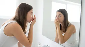 A woman is putting a contact lens into her eye in front of a mirror.