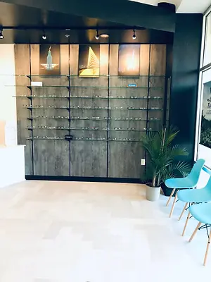 A display case in an optician's office shows a variety of glasses and accessories, with a potted plant and two blue chairs on the side, and a glass wall reflecting images of a sailboat, a fire, and a bottle. The room has a wooden floor and a black wall with a white shelf, and track lights are on the ceiling.