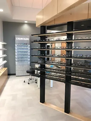 The interior of an eyewear store displays a large display of glasses on shelves, with a Calvin Klein sign visible, a black stool in the corner, and an open door leading to another room. The floor is white tile, and the ceiling is white with a ceiling light.