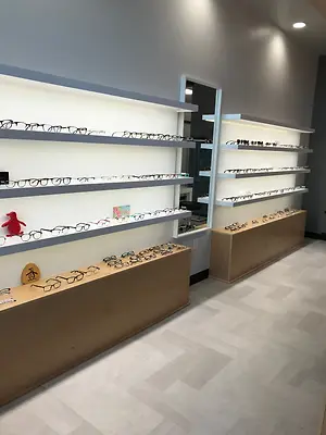 A display of eyeglasses on a wooden shelf with a penguin toy and a mirror in a store with a white tiled floor