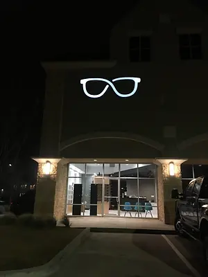 A building with a logo of glasses and a parked vehicle at night, there are chairs inside, and there are lights illuminating the area.