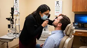 A woman wearing a mask and glasses holds an ophthalmoscope against a man's eye while he sits in a dental chair