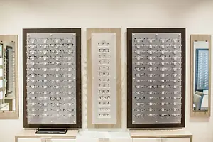 Three glass frames are mounted on a white wall, each containing a different assortment of eyeglasses. The frames are mounted on a white wall with a mirror on the right side and a cabinet below.