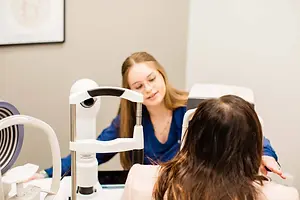 A woman in a blue top is using an eye examination machine on a woman with long hair.