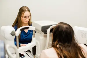 A woman with glasses is looking at a woman sitting in front of her in an eye examination room