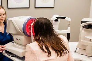 A woman wearing a pink shirt sits in front of an eye scanning machine while a woman in a blue shirt stands behind her and smiles. A wall is behind them with two framed pictures on it.