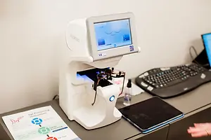 The image shows an advanced device for eye measurement in an office setting, displaying a screen and a paper with eye-pointing instructions.
