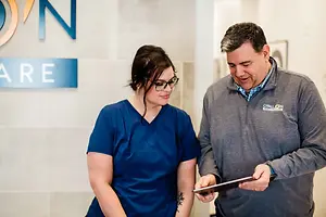 An adult man and woman in blue and gray clothes are looking at a tablet in a medical office