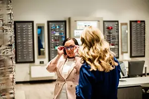 A woman tries on glasses while another woman watches her in a store