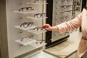A woman wearing a pink long-sleeve shirt is holding a pair of eyeglasses in front of a display of eyeglasses on a white wall