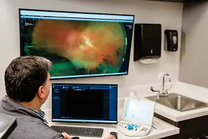 A man is sitting in front of a desk with two monitors and a machine, and he is looking at the monitor on the left side.