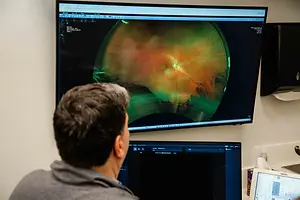 A man is sitting in front of a computer monitor, looking at an eye scan image displayed on the monitor, with another monitor on the desk in front of him.