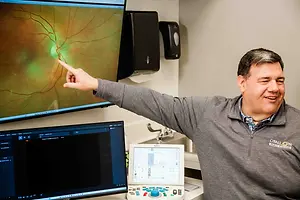 An adult man wearing a gray shirt with the words 'Ottallon' on the front of his shirt is pointing at a monitor that has an image of an eye on it. He is standing in a room that has a sink, monitor, and other equipment on a white counter. He is smiling.