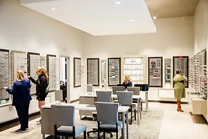 Three women are in an optical shop, looking at frames on the wall. One woman is touching a frame, another is looking at the wall, and the third is standing on the right side.