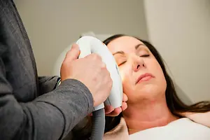 A woman is getting a facial treatment in a room with a white wall, while a man is holding a laser device in front of her face.