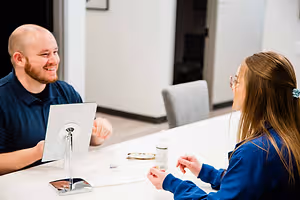 A man and a woman are sitting at a table, with the woman wearing glasses and the man smiling at her, a desk lamp and a bottle are on the table, and a chair and a door are behind them.