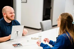A man and a woman are sitting at a table, with the woman wearing glasses and the man smiling at her, a desk lamp and a bottle are on the table, and a chair and a door are behind them.