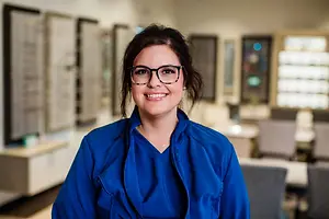 A smiling woman with glasses stands in a room with a white wall, wearing a blue shirt and a necklace, surrounded by picture frames, chairs, and tables.