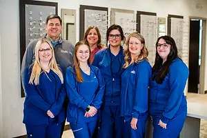 A group of people in blue outfits, possibly employees, are posing for a photo in front of a white wall with framed eyeglasses on it.