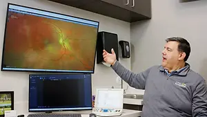 A man is sitting in front of a monitor and pointing to a monitor showing an eye image in a room with cabinets, a sink, and medical equipment