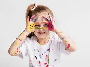 A young girl is smiling and posing with her hands covered in paint, probably for a photo. She has a white t-shirt on, and her hands are covered in different colors of paint. She has a ponytail hairstyle, and her face is slightly dirty with paint. She is probably standing in a room, and the wall behind her is white.
