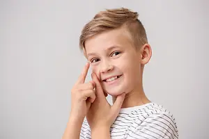 A boy wearing a striped shirt is smiling while making a peace sign gesture with his right hand and touching his right eye with his left hand.