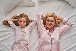 A mother and daughter lying on a bed together, both wearing pink and white striped pajamas and round sunglasses, with a white pillow behind them.