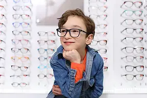 A boy is smiling and posing for a photo while wearing glasses in front of a display of eyeglasses in a store.