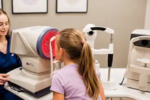 A young girl is having her eyes examined by an optometrist using a medical device in an office setting.