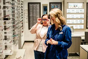 Two women smiling and holding glasses inside a store with shelves full of glasses in the background