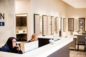 Inside an eye care center, a woman is on a phone call at a reception desk, while another woman is sitting behind the desk, and framed pictures are on the wall.
