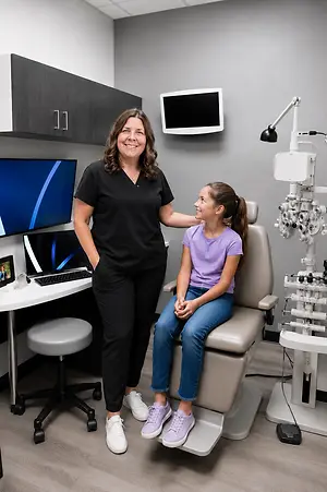 A young girl with pigtails sits in an optometrist's chair while an optometrist stands next to her, both smiling.