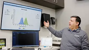 A man wearing a gray sweater stands in a room, gesturing toward a computer monitor displaying a graph and data. Behind him is a sink and a cabinet. There are two other monitors on the desk. There is a picture frame on the left side of the desk, and a white object with a blue button is on the desk.